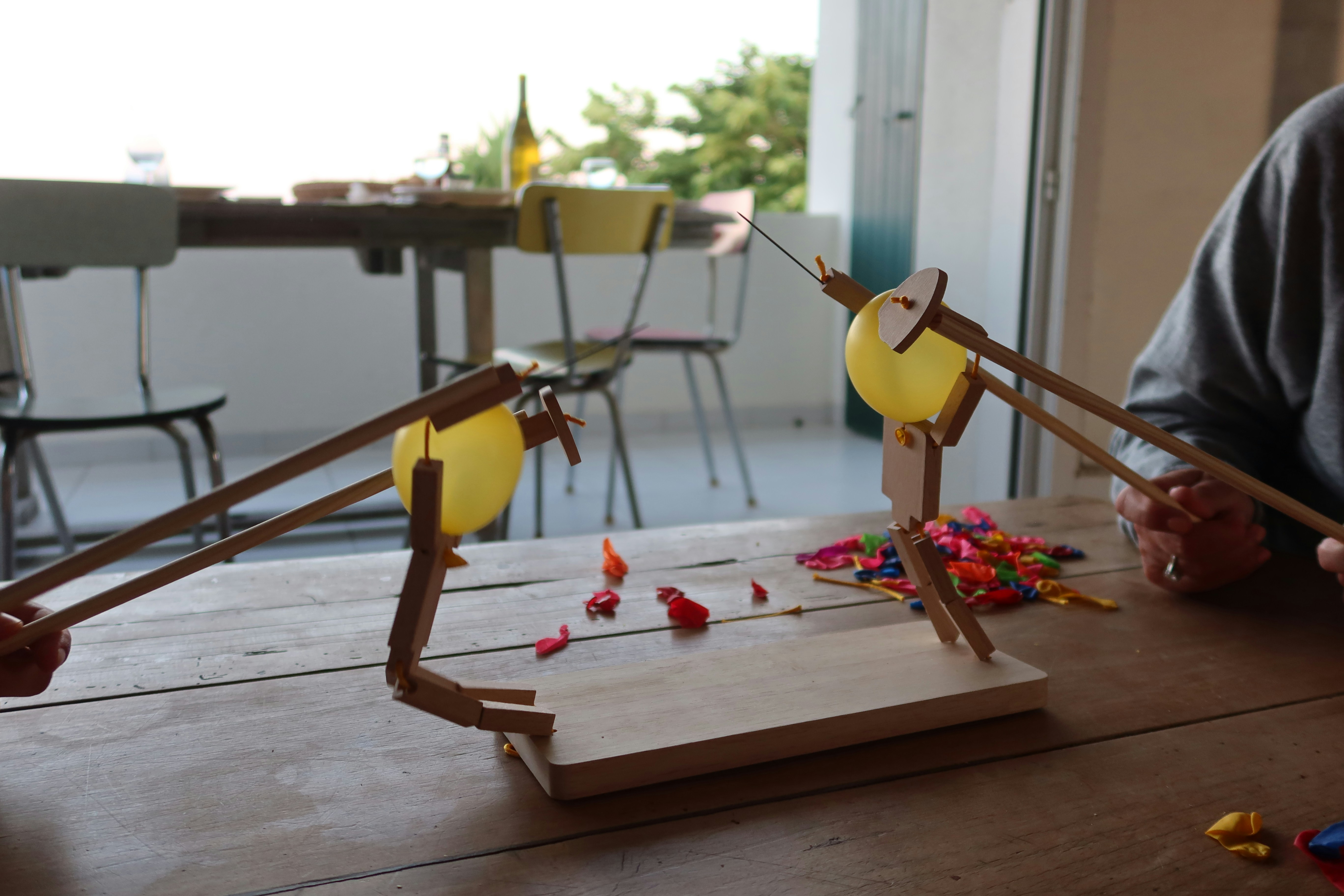 A playful tabletop battle unfolds between two wooden stick‑figure puppets with yellow and blue balloon heads, their skewering rods poised as colorful popped balloon fragments scatter across a rustic wooden table in a bright indoor‑outdoor dining space.